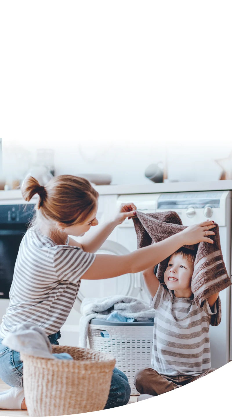 A smiling mother playing with her child in a laundry room, holding a towel over the child’s head, with a washing machine and laundry baskets in the background.